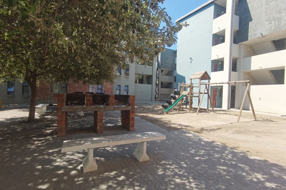 Communal outdoor area featuring a brick braai, concrete bench, and children's playground equipment set within a paved courtyard between apartment blocks.