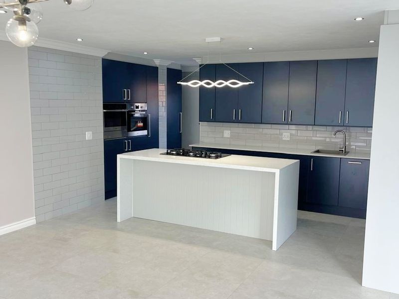 Modern open-plan kitchen featuring dark blue cabinetry, a central island with a gas hob, built-in appliances, and light grey tiled flooring.