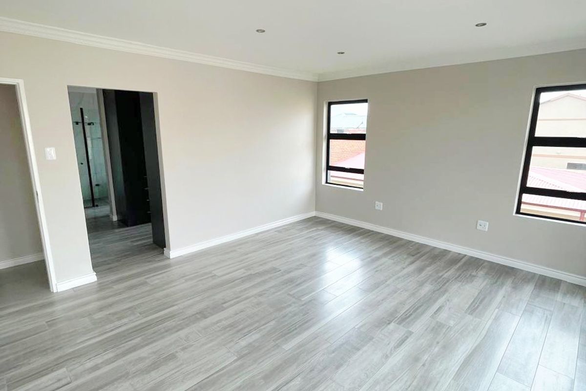A bright, vacant bedroom featuring modern grey laminate flooring, neutral walls, recessed downlighting, and aluminium-framed windows, with an adjacent en-suite bathroom visible.