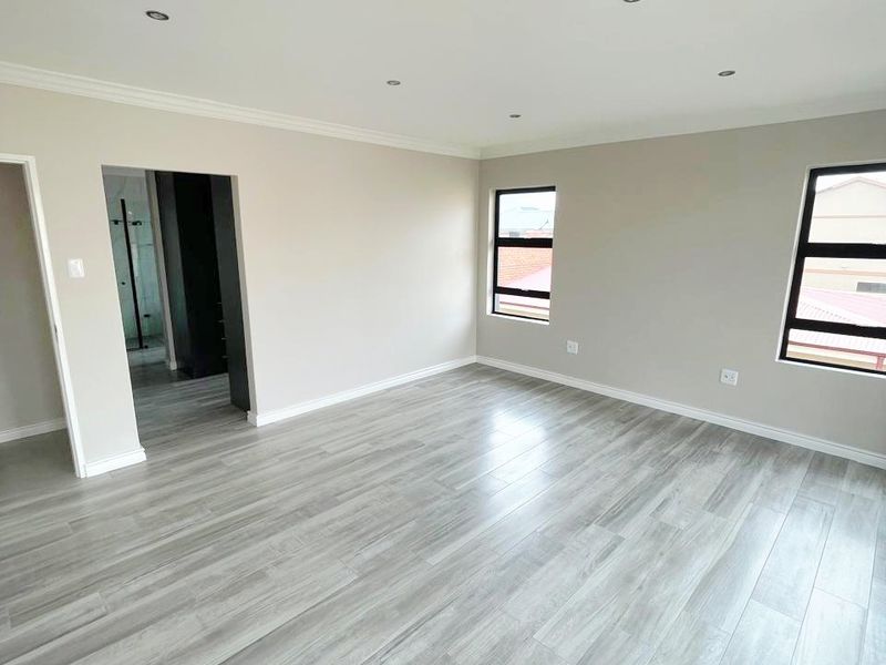 A bright, vacant bedroom featuring modern grey laminate flooring, neutral walls, recessed downlighting, and aluminium-framed windows, with an adjacent en-suite bathroom visible.