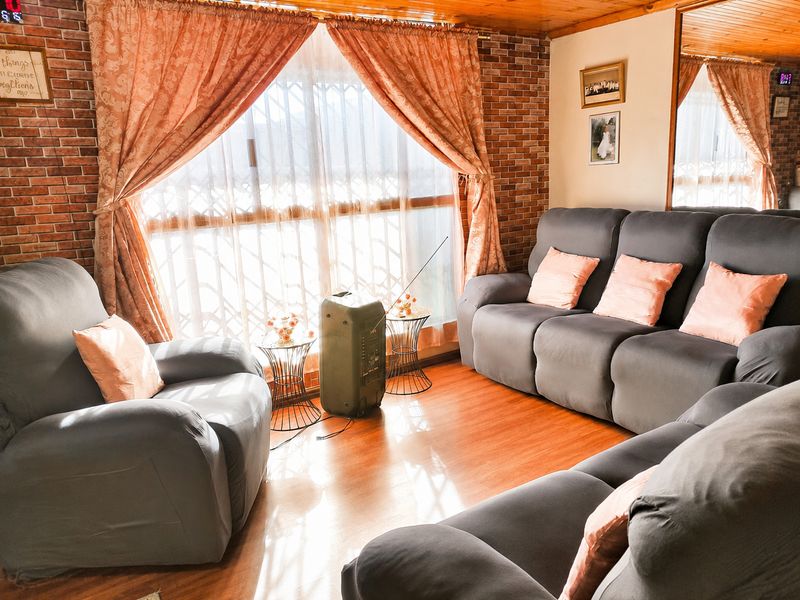 A cozy living room featuring exposed brick walls, warm timber ceiling paneling, and laminate flooring, illuminated by a large window with security gate.
