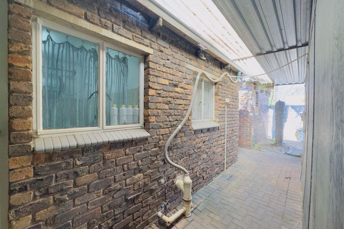 A narrow exterior side passage featuring exposed brick walls, a corrugated metal roof covering, and paved flooring with visible external plumbing infrastructure.