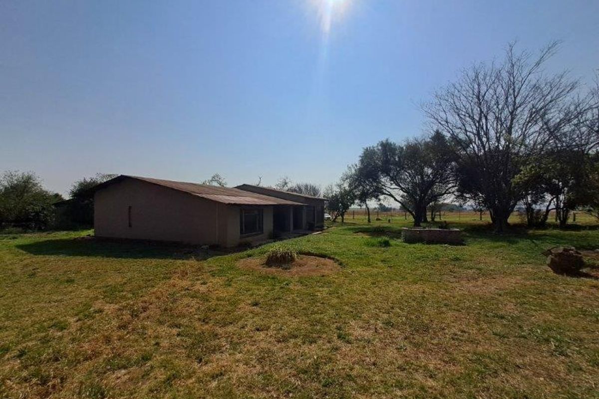 Rear view of a farm dwelling featuring a corrugated metal roof, a concrete patio area, and a stone well set within a large garden.