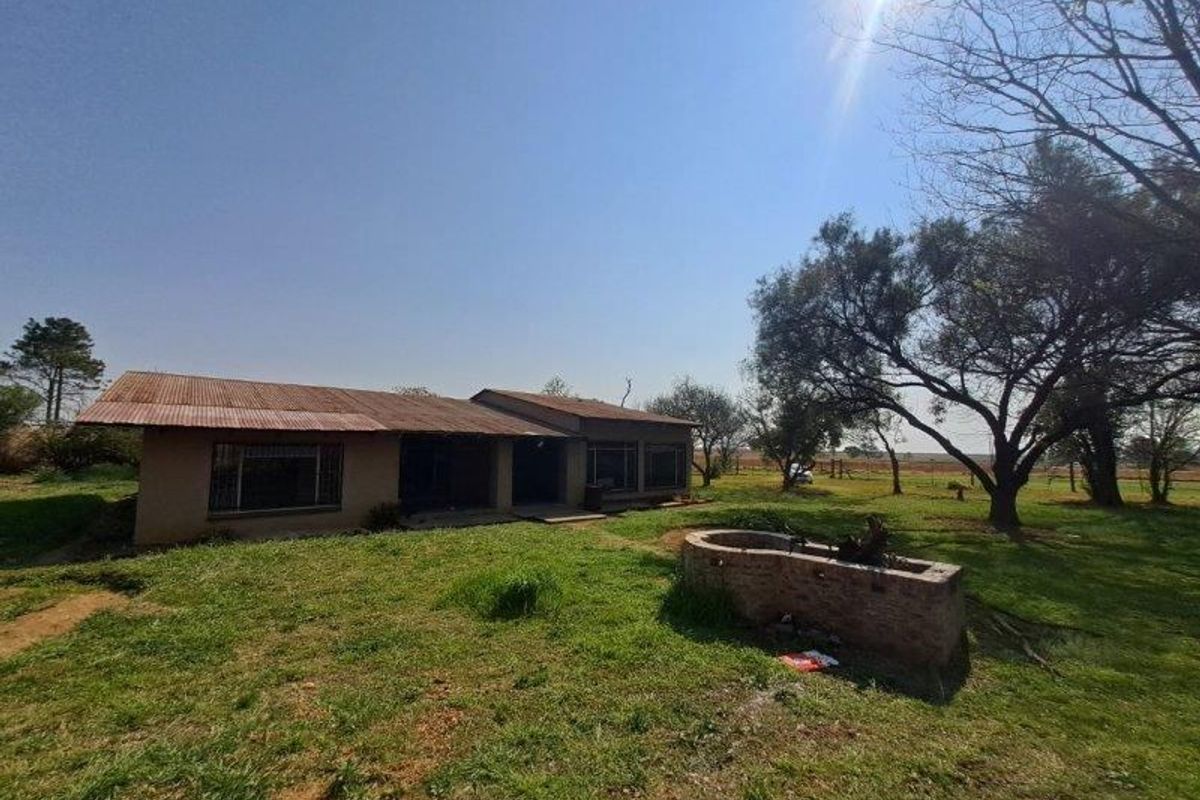 Rear view of a farm dwelling featuring a corrugated metal roof, brick construction, and a brick water feature set within a large rural garden.