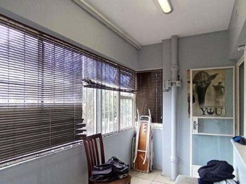 Enclosed balcony area featuring tiled flooring, large sliding windows with venetian blinds, and exposed utility piping.