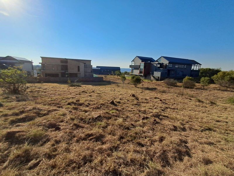 A view of a vacant residential plot featuring natural dry grass, wire fencing, and surrounding modern residential properties.