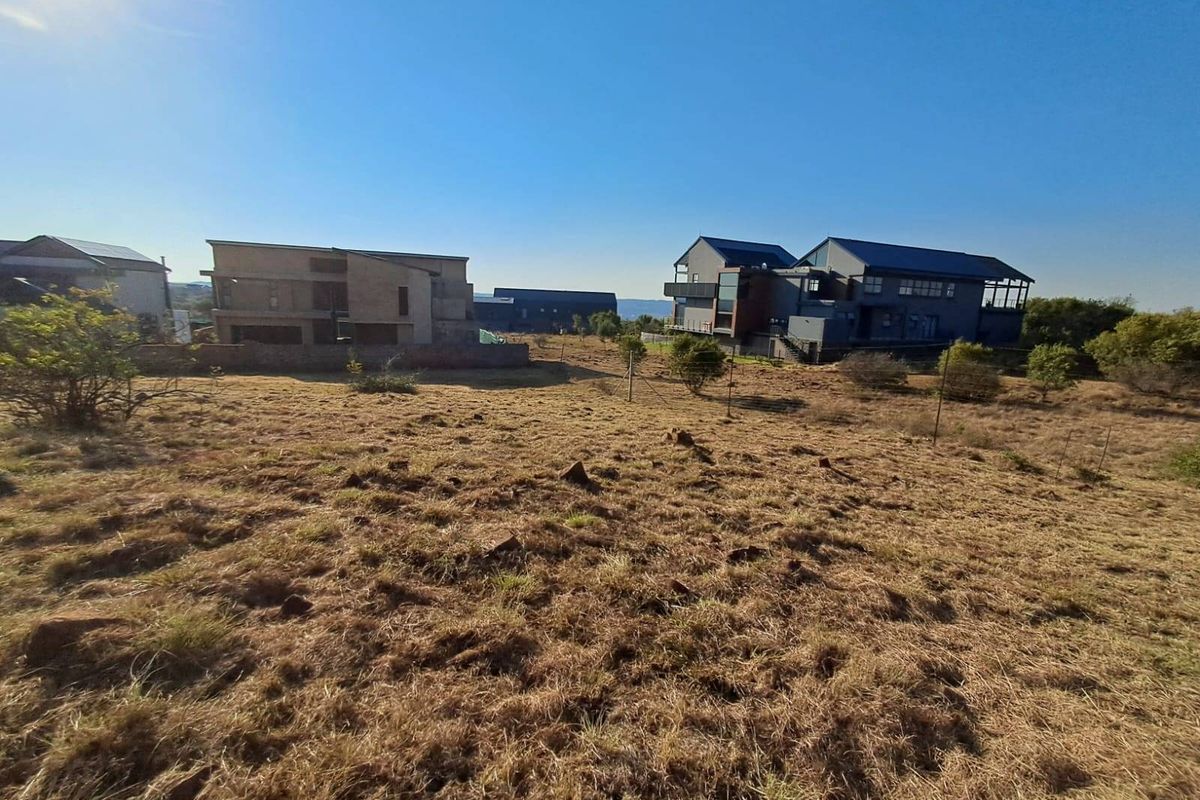 A view of a vacant residential plot featuring natural dry grass, wire fencing, and surrounding modern residential properties.