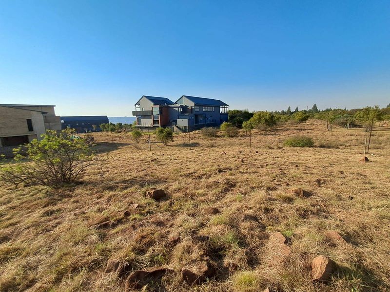 A view of a vacant residential plot featuring natural dry grass, scattered rocks, and surrounding residential properties in the background.