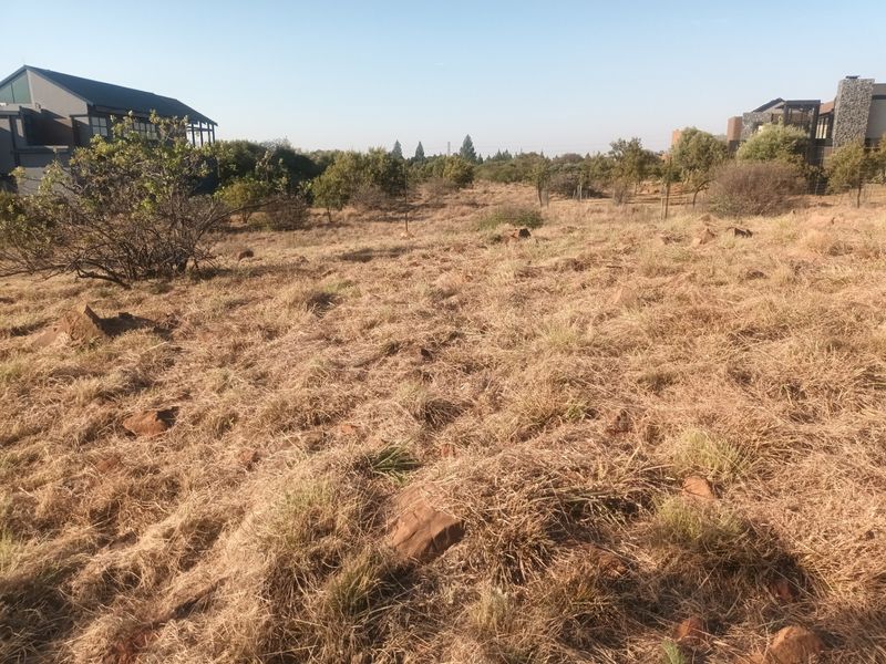 A view of a vacant residential plot featuring natural dry grass, rocky terrain, and surrounding established residential properties.