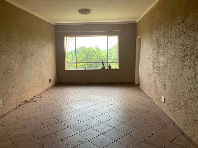 An empty, neutrally-toned living room featuring tiled flooring, textured walls, and a large window providing natural light and a view of the greenery outside.