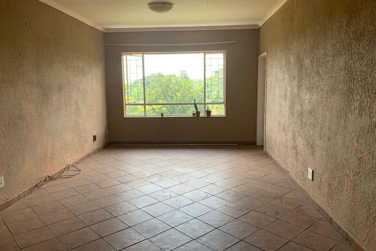 An empty, neutrally-toned living room featuring tiled flooring, textured walls, and a large window providing natural light and a view of the greenery outside.