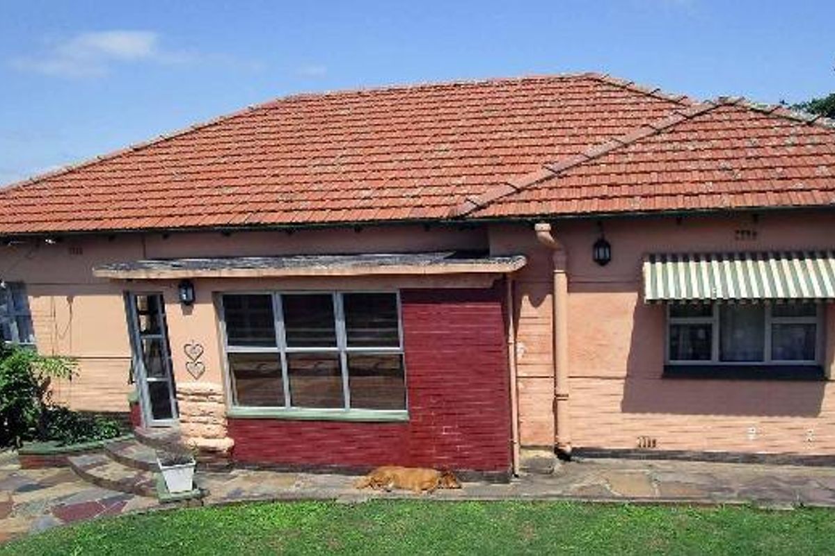 Rear view of a single-story house featuring a terracotta tiled roof, brick facade, metal-framed windows with an awning, and a stone-paved patio area.