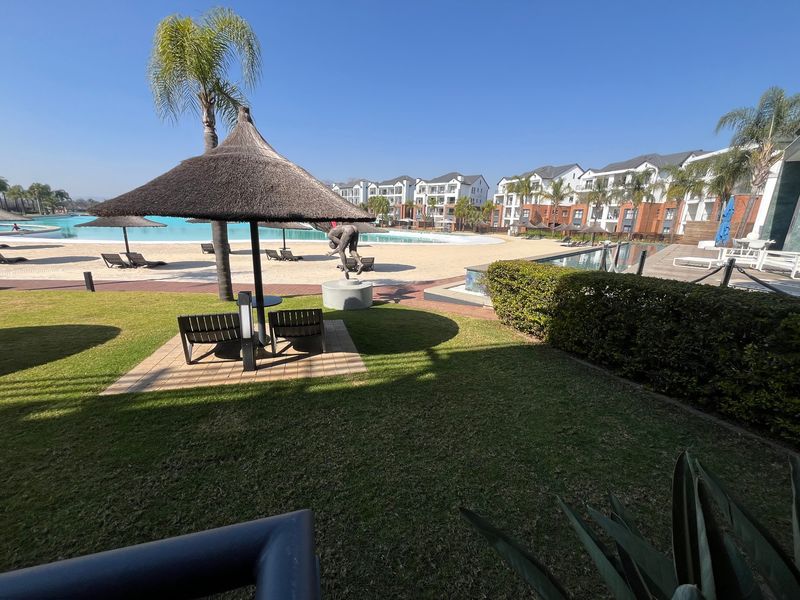 A well-maintained communal pool area featuring a sandy beach entry, thatched umbrellas, and landscaped gardens within a modern apartment complex.