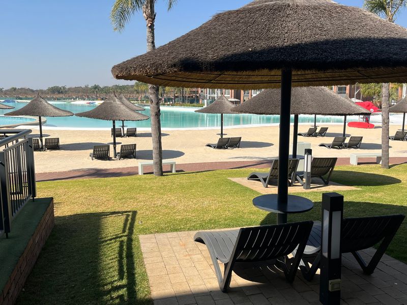 A well-maintained communal pool area featuring a sandy beach entry, thatched umbrellas, paved patio space, and manicured lawns.