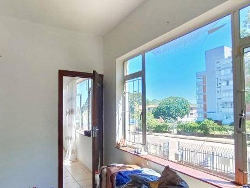 A bright, empty living area featuring large metal-framed windows with security bars, offering an urban street view and natural light.