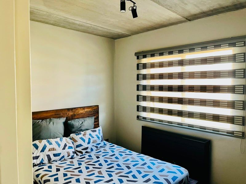 A cozy bedroom featuring an exposed concrete ceiling, modern track lighting, and zebra roller blinds for adjustable natural light.