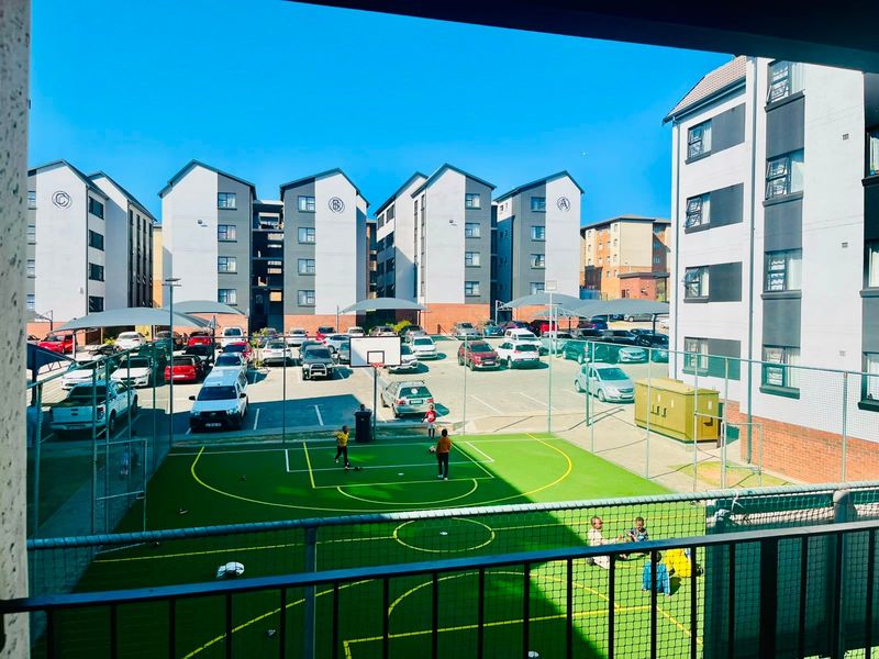Elevated view overlooking a residential apartment complex featuring a fenced sports court, paved parking area with shade cloth carports, and multi-story building blocks.