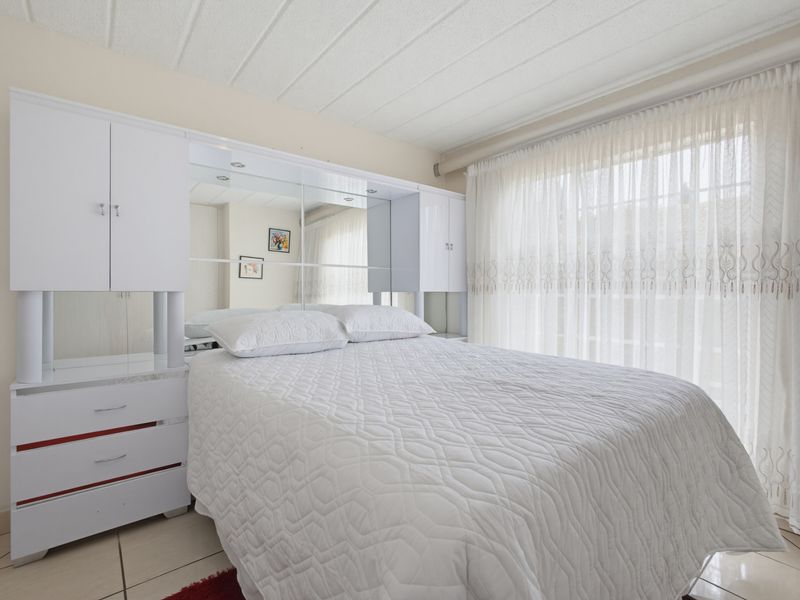 A bedroom featuring built-in white cabinetry with a mirrored headboard, tiled flooring, and large windows with sheer curtains.