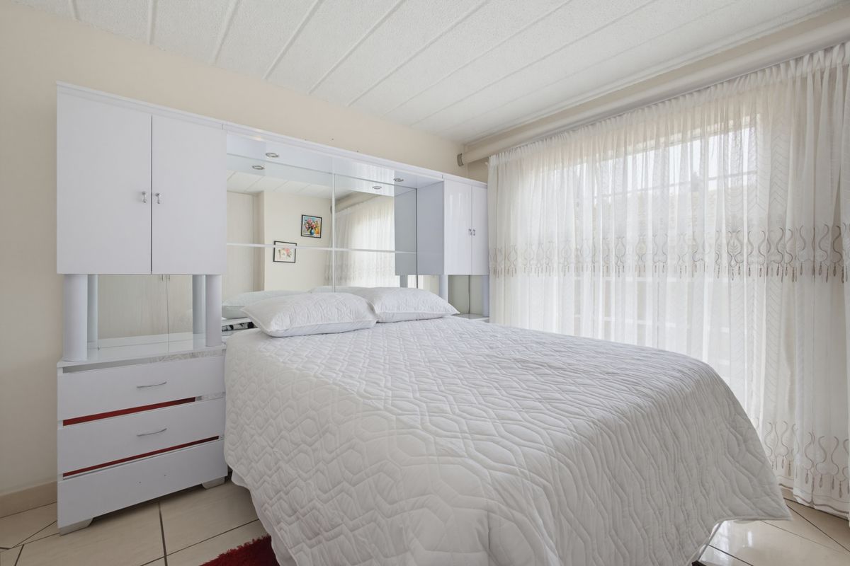 A bedroom featuring built-in white cabinetry with a mirrored headboard, tiled flooring, and large windows with sheer curtains.