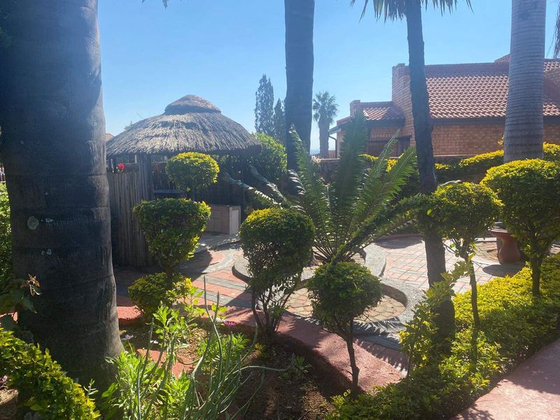 A landscaped garden area featuring a traditional thatched lapa and paved patio space, surrounded by mature trees and established shrubbery.