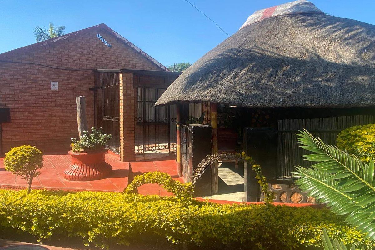 Rear exterior view featuring a traditional thatched lapa, brick building facade, and a paved patio area set within a landscaped garden.