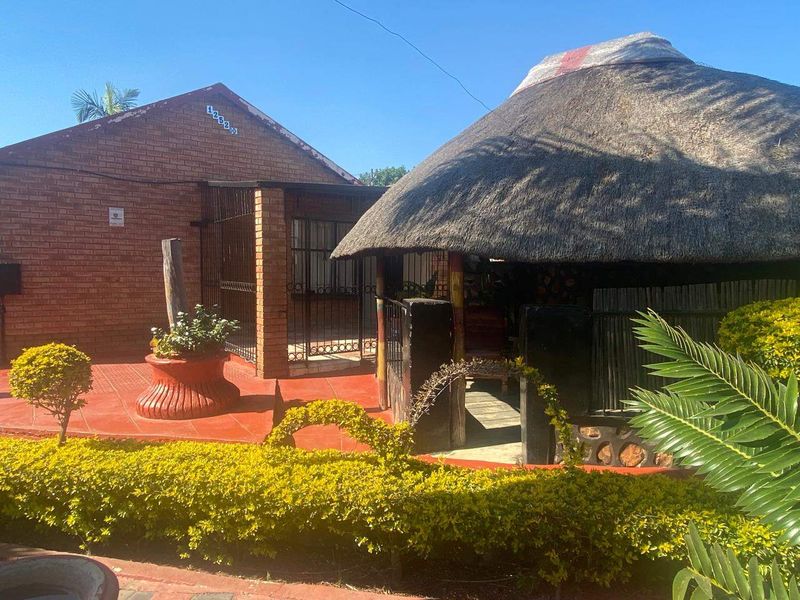 Rear exterior view featuring a traditional thatched lapa, brick building facade, and a paved patio area set within a landscaped garden.