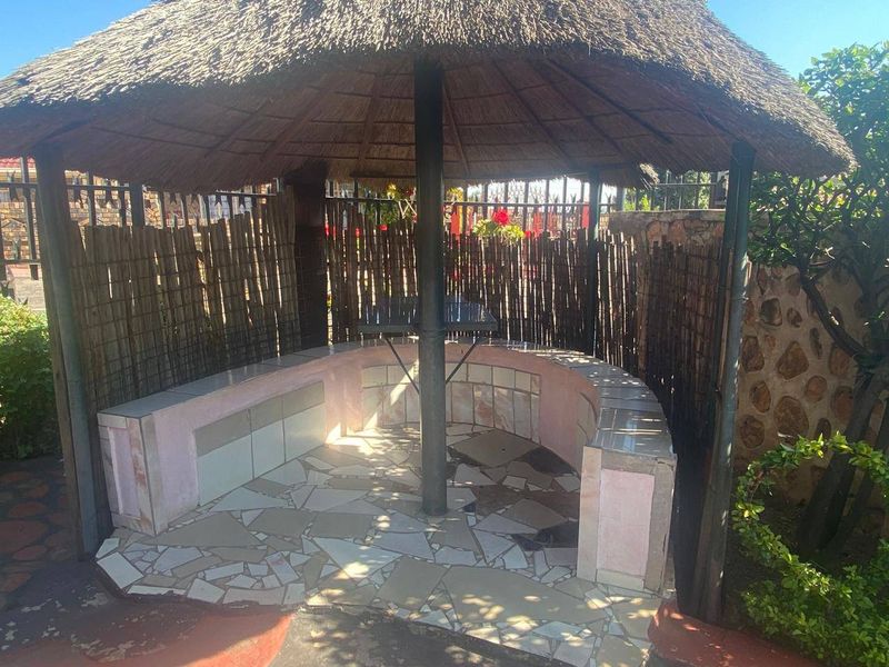 Outdoor thatched lapa area featuring built-in curved seating and stone-tiled flooring, providing a rustic entertainment space in the garden.