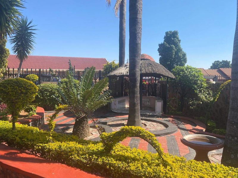 A well-established garden featuring a thatched gazebo, paved circular pathways, mature palm trees, and stone retaining walls.