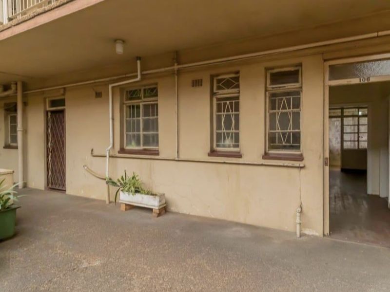 A covered patio area featuring concrete flooring, security gates, and steel-framed windows, showing signs of wear and requiring maintenance.