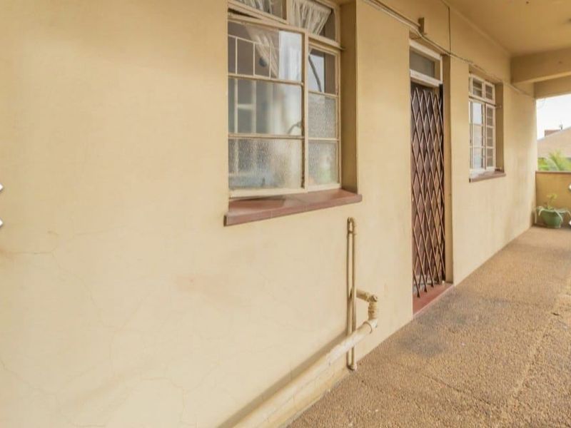 A covered balcony area featuring steel-framed windows, a security gate, and textured concrete flooring, showing signs of wear and requiring cosmetic maintenance.