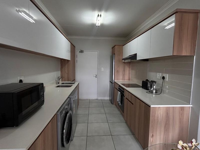 Modern galley-style kitchen featuring wood-grain cabinetry, white high-gloss upper cabinets, stone countertops, integrated appliances, and neutral tiled flooring.