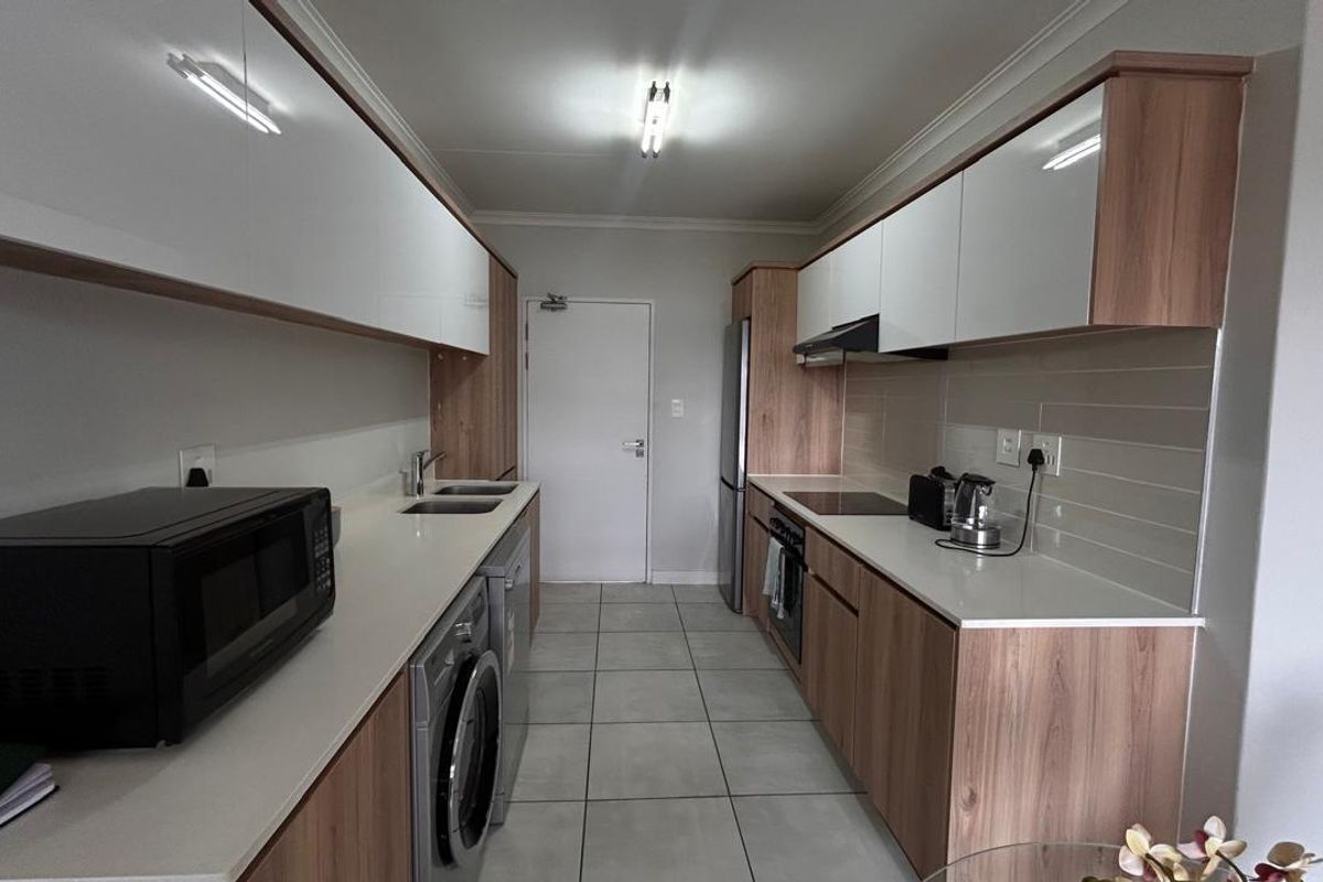Modern galley-style kitchen featuring wood-grain cabinetry, white high-gloss upper cabinets, stone countertops, integrated appliances, and neutral tiled flooring.