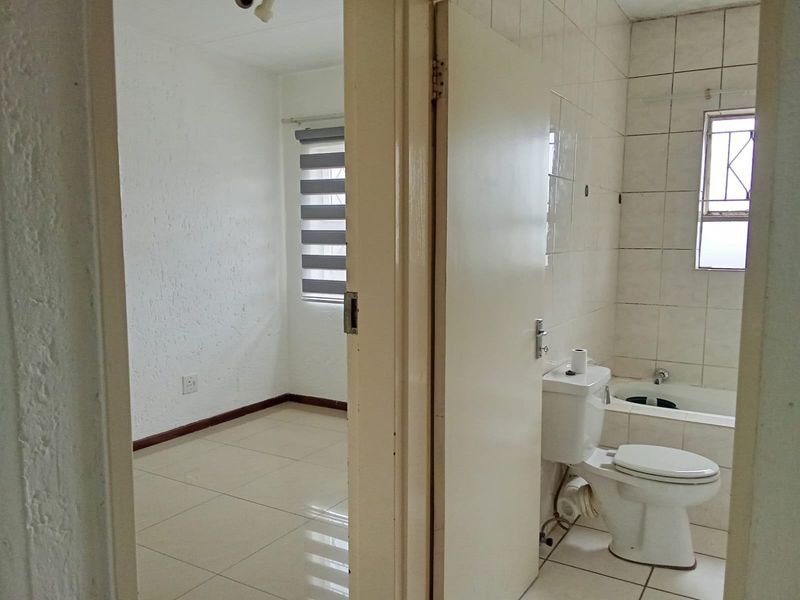 A dated bathroom featuring tiled walls and flooring, a built-in bathtub, and a standard toilet, adjacent to an empty bedroom space.