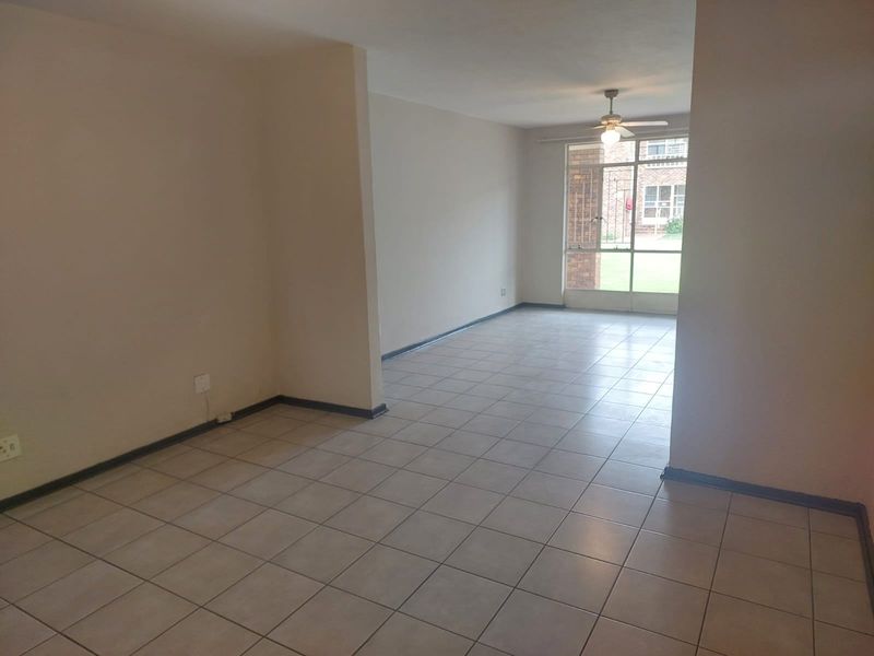 A spacious, empty living area featuring neutral tiled flooring, a ceiling fan, and large windows providing natural light and garden views.