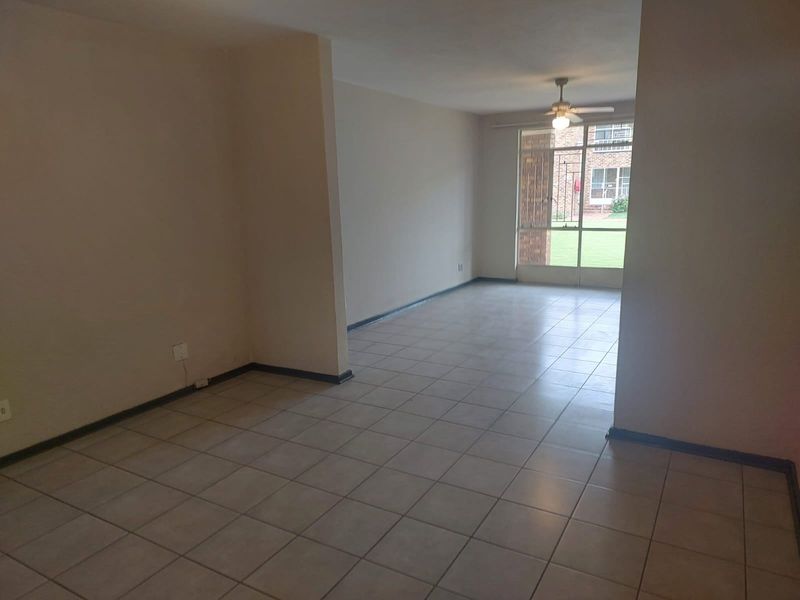 An empty, open-plan living area featuring neutral tiled flooring, a ceiling fan, and large windows providing natural light and garden views.