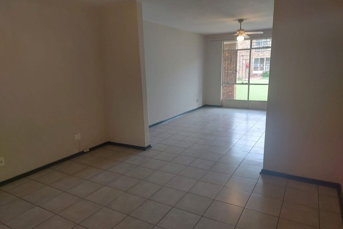 An empty, open-plan living area featuring neutral tiled flooring, a ceiling fan, and large windows providing natural light and garden views.