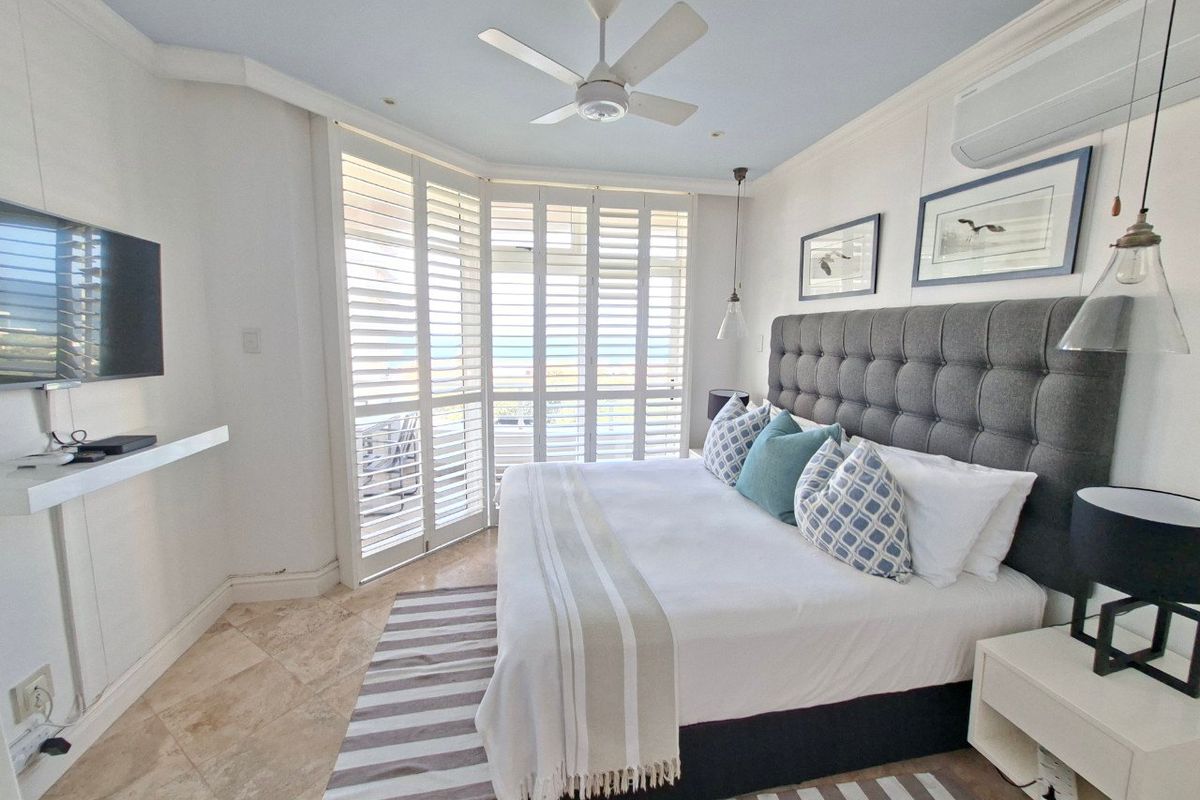 A well-maintained bedroom featuring stone flooring, a ceiling fan, split-system air conditioning, and plantation shutters opening to a balcony view.