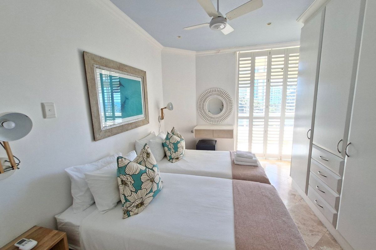 A bright, well-maintained bedroom featuring built-in cupboards, a ceiling fan, and plantation shutters leading to a balcony with natural light.