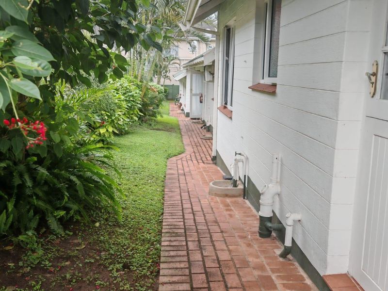 A side view of the property featuring a brick paved walkway, landscaped garden, and external plumbing fixtures along the white exterior wall.