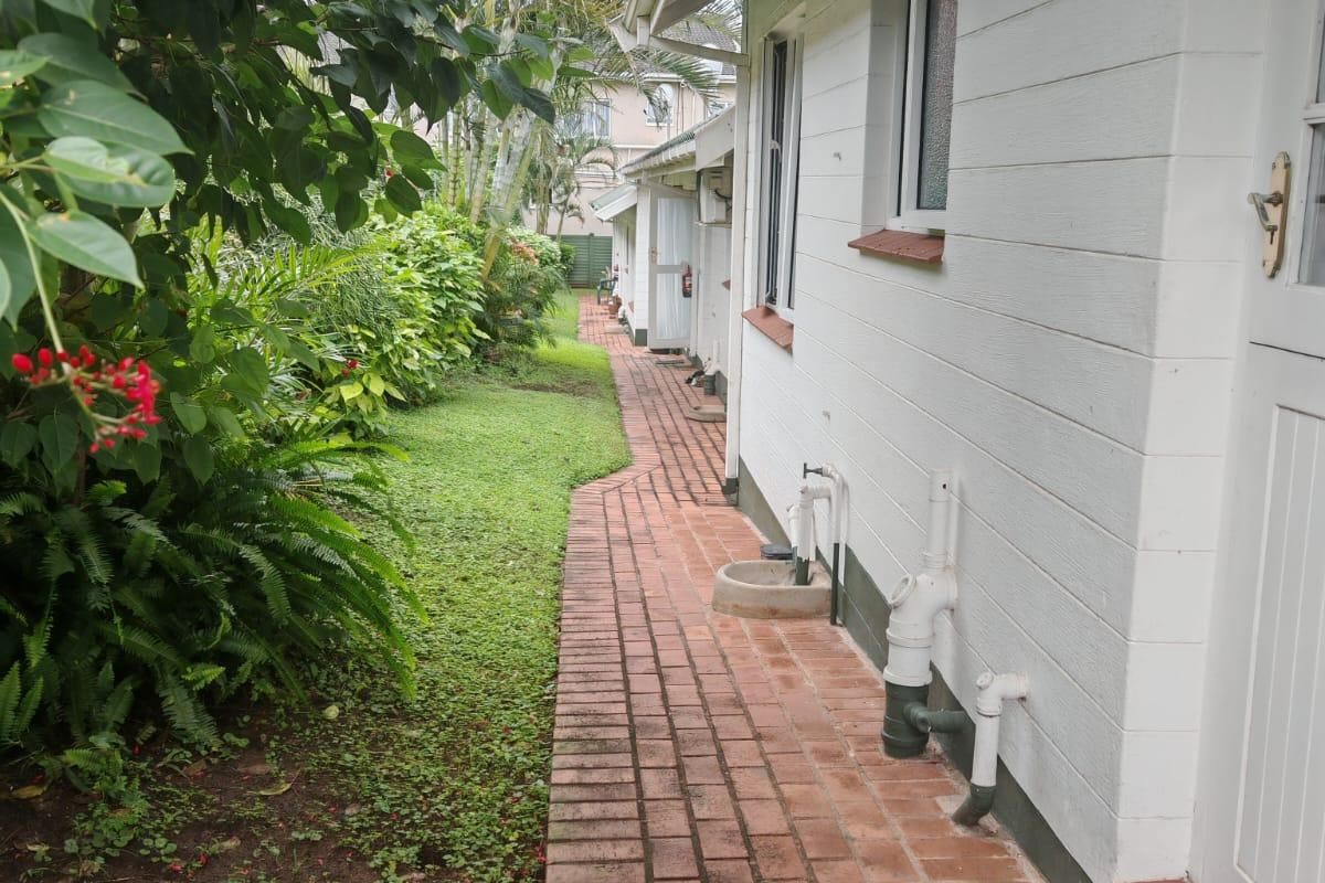 A side view of the property featuring a brick paved walkway, landscaped garden, and external plumbing fixtures along the white exterior wall.