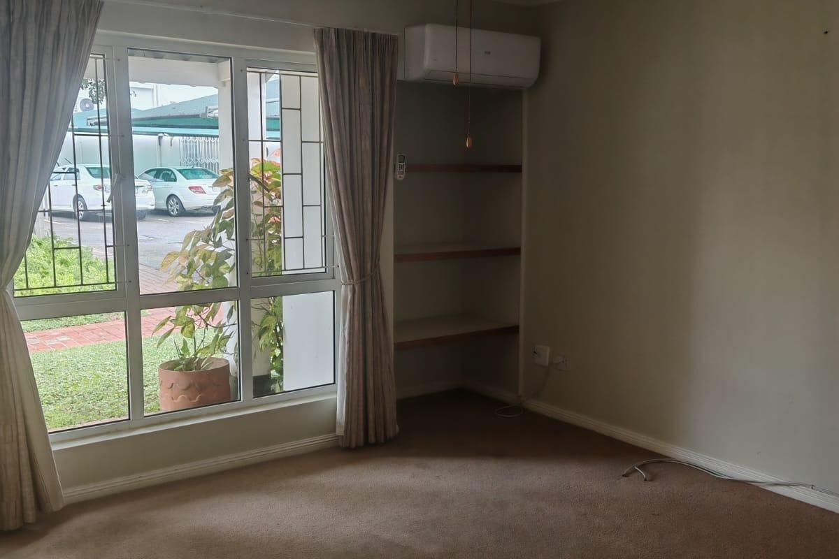 A spacious, carpeted living area featuring a ceiling fan, wall-mounted air conditioning unit, and built-in shelving, with large windows overlooking the exterior.