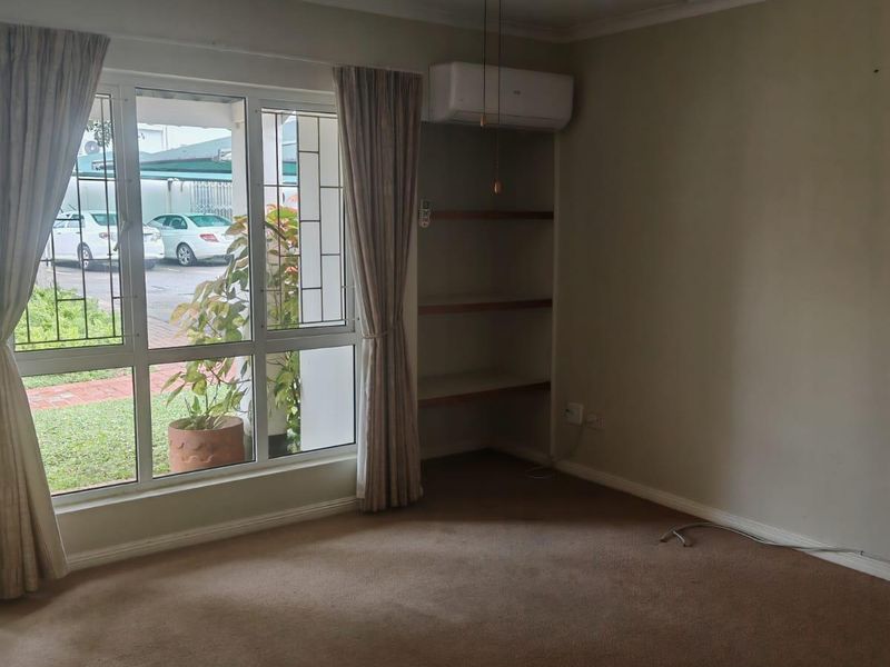 A spacious, carpeted living area featuring a ceiling fan, wall-mounted air conditioning unit, and built-in shelving, with large windows overlooking the exterior.