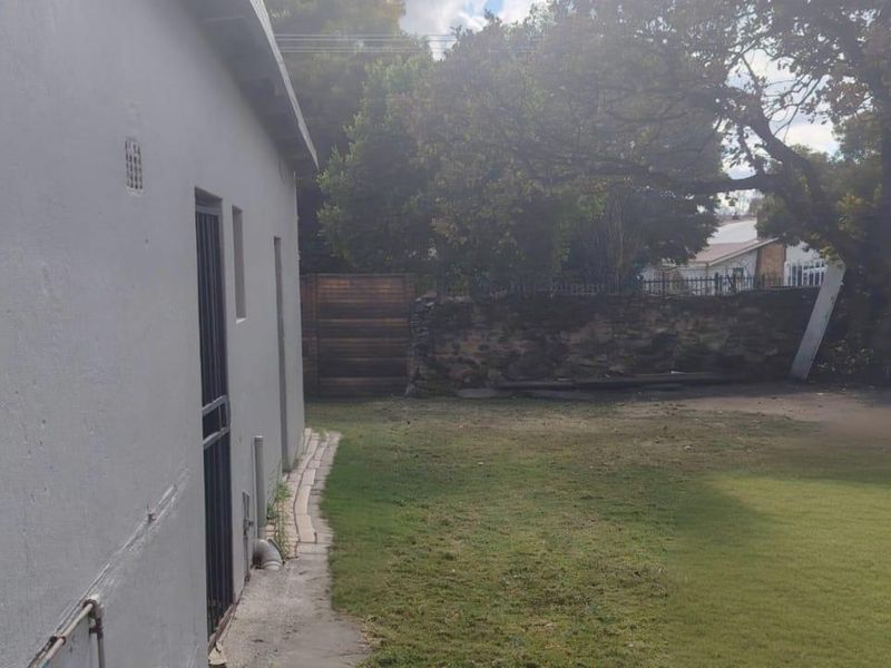 A view of the rear exterior of the property featuring a lawn, stone boundary wall, and paved walkway along the side of the house.