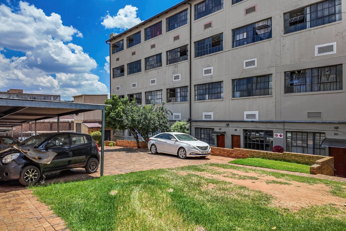 Rear exterior view of an industrial-style apartment building featuring a paved parking area, carport, and communal lawn space.