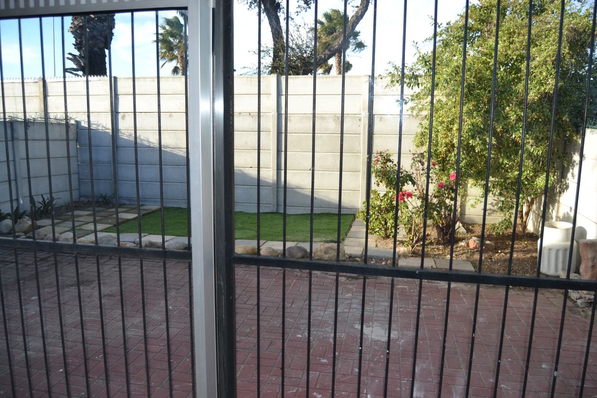 A private rear garden area featuring a paved patio, small lawn, and secure perimeter walling, viewed through a metal security gate.