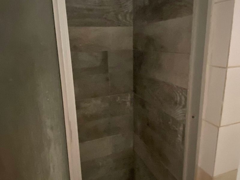 A compact shower stall featuring wood-look wall tiles and a mosaic tiled floor, requiring some maintenance to the frame and grout.