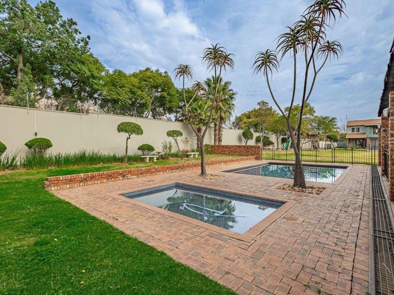 A well-maintained outdoor pool area featuring dual swimming pools, brick paving, and established landscaping enclosed by a secure perimeter wall.