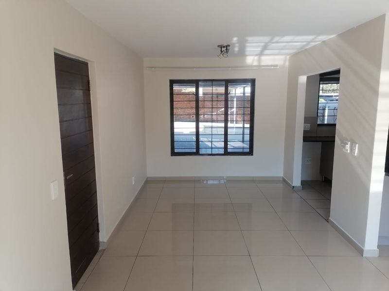 A bright, empty open-plan living area featuring neutral tiled flooring, clean walls, and a window with security bars, leading into an adjacent kitchen space.