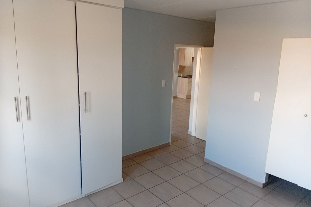 A clean, empty bedroom featuring neutral wall tones, tiled flooring, and built-in cupboards, with a view through to the kitchen area.