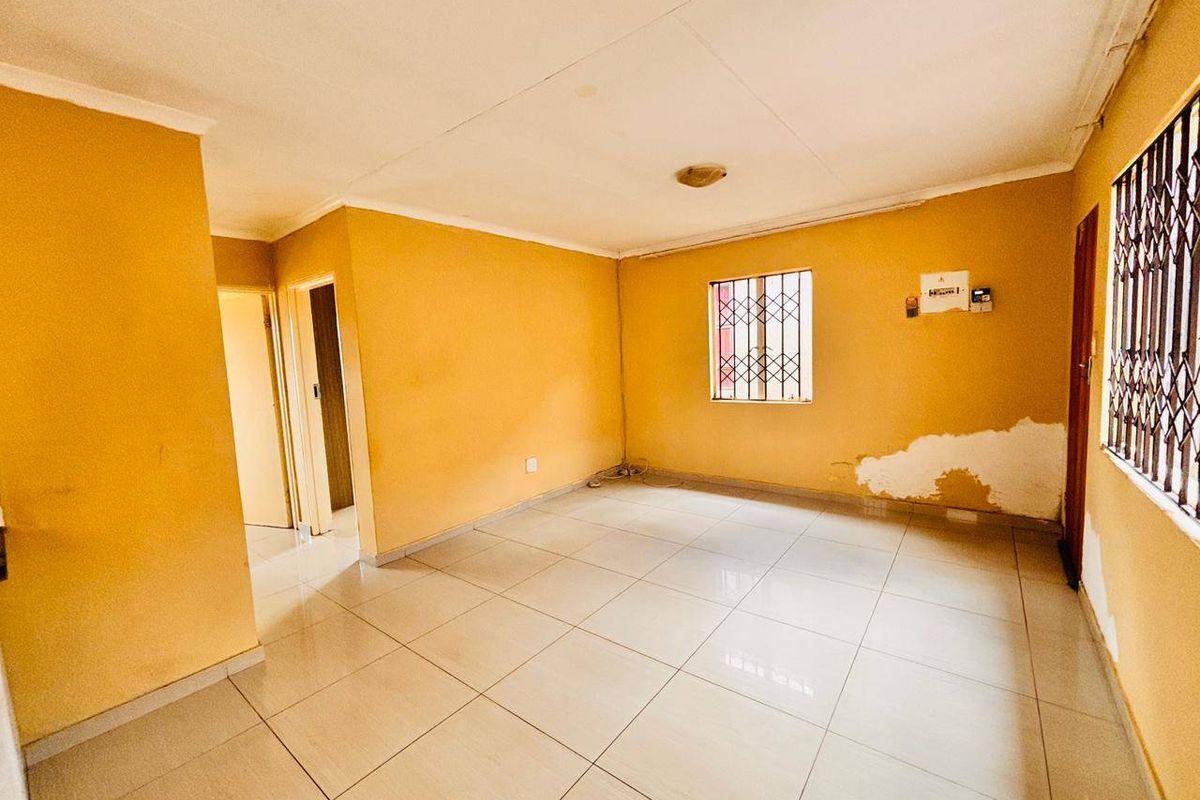 An empty living room featuring tiled flooring and security bars on the windows, currently showing signs of wall damage requiring maintenance.